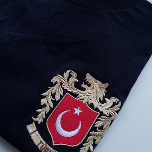 Op de afbeelding: Zwart T-shirt met een geborduurd wapenschild met een gouden leeuw, een rood schild met een witte halve maan en ster, en het woord "TÜRKIYE" in gouden letters.
