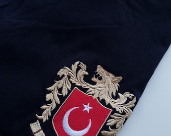 Może przedstawiać: Czarny T-shirt z haftowanym herbem przedstawiającym złotego lwa, czerwoną tarczę z białym półksiężycem i gwiazdą oraz napis "TÜRKIYE" złotymi literami.