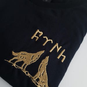 Op de afbeelding: Zwart T-shirt met een gouden geborduurd ontwerp van twee wolven die naar de maan huilen. De tekst "RYNH" is ook in goud geborduurd.