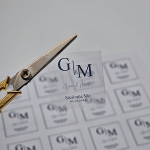 Könnte beinhalten: Ein transparenter Aufkleber mit den Initialen "G|M" und dem Text "Gızem & Murat" und "Sözümüz Söz 02.03.2024". Der Aufkleber befindet sich auf einem Blatt Papier mit mehreren Kopien desselben Aufklebers.