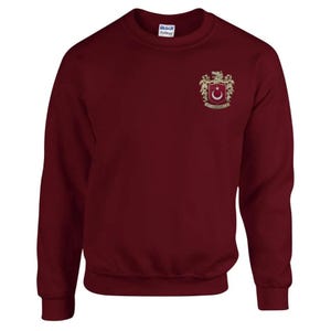 Op de afbeelding: Een bordeauxrood sweatshirt met een geborduurd gouden wapenschild met een halve maan en een ster, omringd door twee leeuwen.