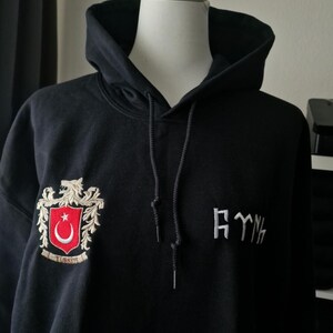 Op de afbeelding: Zwarte hoodie met een trekkoord, met een geborduurd wapen met een rode maan en ster, en het woord "TURKIYE". De hoodie heeft ook geborduurde tekst in een gestileerd lettertype.