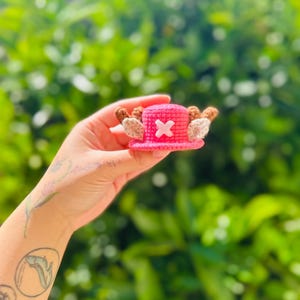 May include: A miniature crocheted pink hat featuring a white cross emblem on the front and two brown textured antlers extending from the sides. The small accessory is crafted with a visible yarn stitch pattern and has a rounded top with a brim.