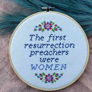 Op de afbeelding: Een witte borduurring met een houten frame. De borduurwerk leest "The first resurrection preachers were WOMEN" met een bloemenontwerp boven en onder de tekst.