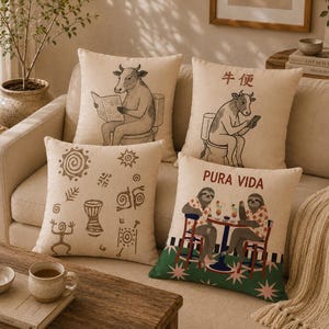 May include: Four square throw pillows with beige fabric covers. One features a cow reading a newspaper on a toilet, another shows a cow on a toilet with Chinese characters, one has brown tribal-style symbols, and one depicts two sloths with the text Pura Vida.