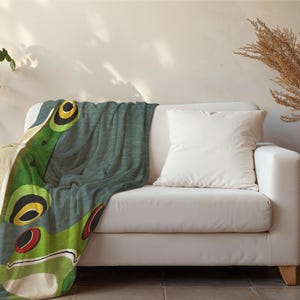 Puede incluir: Una manta suave de color verde azulado y verde con un diseño de rana. La manta está colocada sobre un sofá blanco con un cojín blanco.