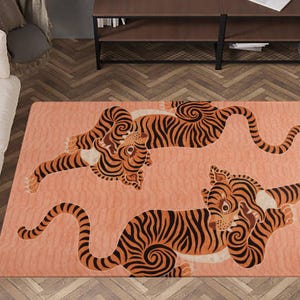 Könnte beinhalten: Ein rechteckiger Teppich mit pfirsichfarbenem Hintergrund zeigt drei stilisierte Tiger in Orange und Schwarz. Die Tiger befinden sich in verschiedenen Posen, wobei ihre Körper und Schwänze gekrümmt sind. Der Teppich liegt auf einem Holzboden.