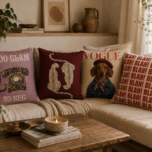 May include: Four square scatter cushions arranged on a sofa. The first is mauve with a leopard print rotary phone and the text TOO GLAM TO BEG. The second is dark red with two gold cheetahs. The third is beige with a dog wearing a beret and the text VOGUE. The fourth is cream with the word BLAH repeated in red.