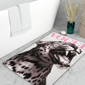 Könnte beinhalten: Rechteckige Badematte mit einem schwarz-weißen Leopardenmuster, das einen geöffneten Mund und Zähne zeigt. Das Wort "VOGUE" ist rosa aufgedruckt. Die Matte liegt auf einem gefliesten Boden in der Nähe einer weißen Badewanne.