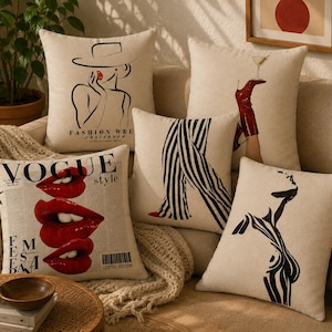 May include: A set of five square throw pillows with beige covers featuring black and red graphic illustrations. Designs include a woman in a hat, a leg in a red boot holding a martini, black and white striped trousers, a silhouette, and red lips with the text VOGUE style.