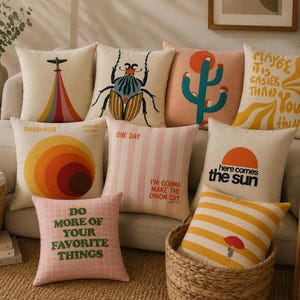 May include: A collection of nine square throw pillows featuring various graphic designs, including a beetle, a cactus, a plane with a rainbow trail, and text such as "here comes the sun," "maybe it is easier than you think," and "do more of your favorite things."