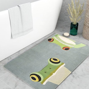 May include: A gray bath mat with a green frog design. The frog has yellow eyes and red spots. The mat is made of a soft, absorbent material.