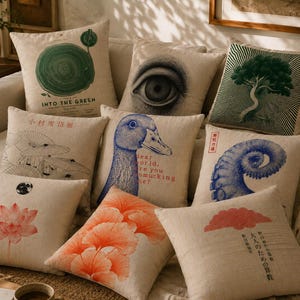 May include: A collection of nine square throw pillows with beige linen-textured covers, each featuring distinct graphic prints including a green tree ring, a human eye, a dark green tree, umbrellas, a blue duck, a blue tentacle, a pink lotus, orange ginkgo leaves, and a red cloud with Japanese text.