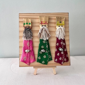 May include: Three Wise Men Christmas ornament, made of fabric and wood. The figures are dressed in pink, green, and burgundy robes with gold accents. Each figure has a crown and a beard.