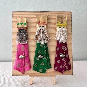 May include: Three Wise Men figurines on a wooden easel. Each figurine is made of fabric and has a gold crown, a white beard, and a colourful robe. The robes are pink, green, and burgundy with gold accents.