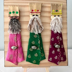 May include: Three paper mache figures of the Three Wise Men, each with a crown, beard, and robe. The figures are mounted on a wooden board with a small easel.