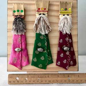 May include: Three Wise Men figures made of wood and fabric. Each figure has a gold crown, a white beard, and a colourful robe. The figures are arranged in a row on a wooden background.