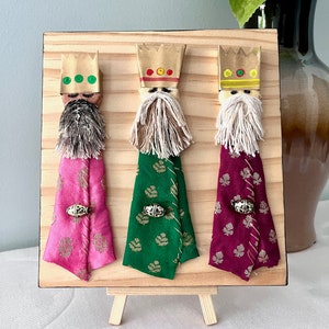 May include: Three Wise Men figurines on a wooden easel. Each figurine is made of fabric and has a gold crown, a white beard, and a colourful robe. The figurines are arranged in a row, with the pink robe on the left, the green robe in the middle, and the burgundy robe on the right.