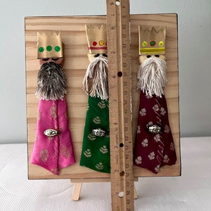 May include: Three Wise Men figurines on a wooden easel. Each figurine is wearing a crown, a robe, and a beard. The robes are pink, green, and burgundy with gold accents. The figurines are approximately 10 inches tall.