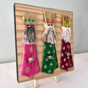 May include: Three Wise Men figurines on a wooden stand. Each figurine is made of fabric and has a gold crown, a white beard, and a different coloured robe: pink, green, and burgundy.