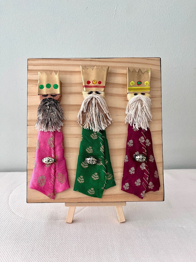 May include: Three Wise Men figurines on a wooden easel. Each figurine is made of fabric and has a gold crown, a white beard, and a colourful robe. The robes are pink, green, and burgundy with gold accents.
