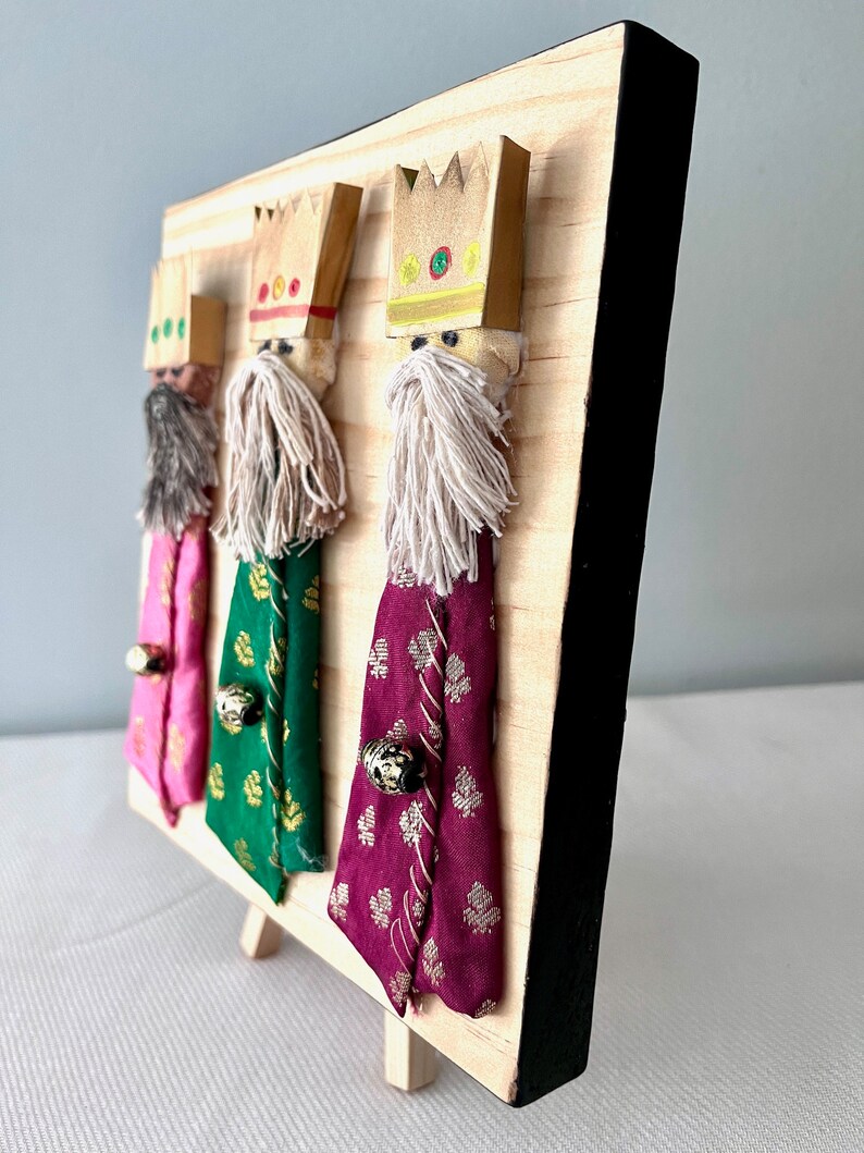 May include: Three wooden figures of the three wise men, each wearing a crown and a robe. The figures are standing on a wooden base with a black frame.