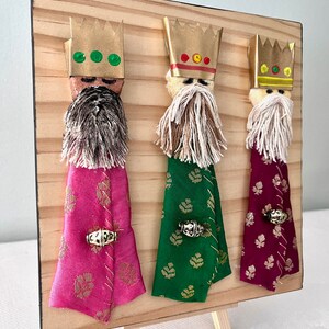May include: Three Wise Men figurines on a wooden easel. Each figurine has a gold crown, a white beard, and a colourful robe. The robes are pink, green, and burgundy.