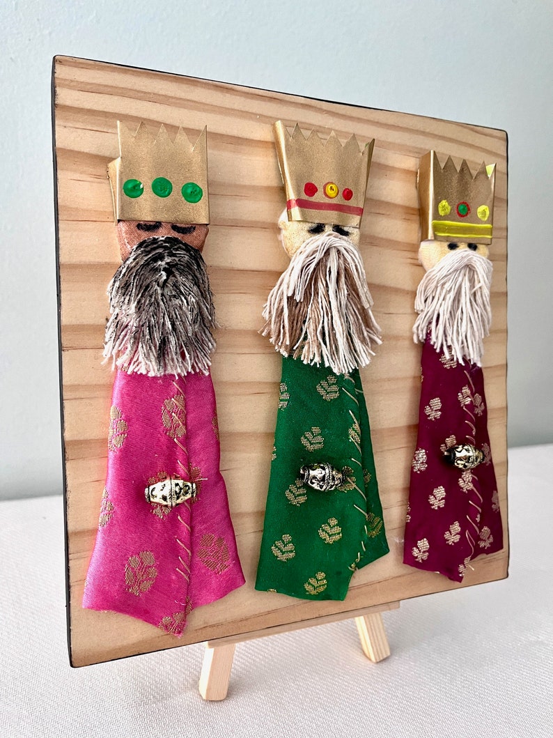 May include: Three Wise Men figurines on a wooden easel. Each figurine has a gold crown, a white beard, and a colourful robe. The robes are pink, green, and burgundy.