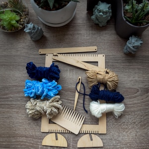 May include: A wooden weaving loom kit with various colored yarn bundles, a comb, and a needle. The yarn colors include navy blue, light blue, beige, and white. The kit is arranged on a wooden surface, with small decorative statues and potted plants in the background.