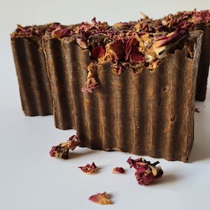 Pode incluir: Três barras de sabão marrons com pétalos de rosa secos no topo. O sabão está sobre uma superfície branca.
