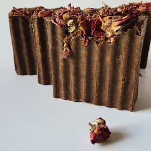 May include: Two brown bars of soap with dried rose petals on top. The soap is textured and has a rustic look. A single dried rose petal is in the foreground.