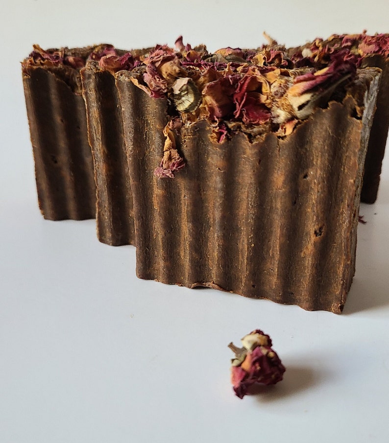 May include: Two brown bars of soap with dried rose petals on top. The soap is textured and has a rustic look. A single dried rose petal is in the foreground.