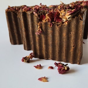 May include: Two brown bars of soap with dried rose petals on top. The soap is on a white surface.