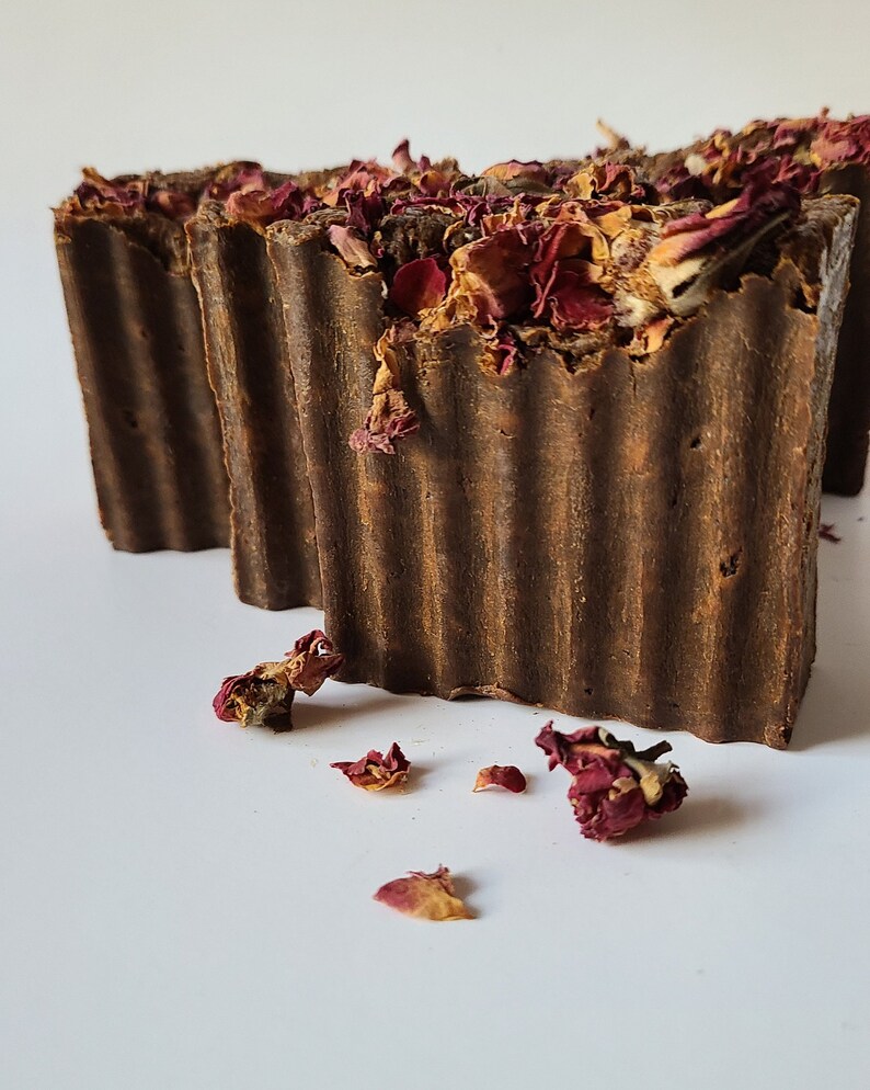 May include: Two brown bars of soap with dried rose petals on top. The soap is on a white surface.