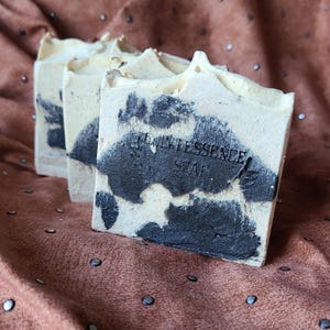 May include: Three bars of handmade soap with a creamy white and dark gray pattern. The soap has a textured surface and the text "QUINTESSENCE SOAP" is stamped on the front. The soaps are arranged on a brown fabric background.