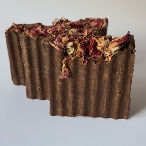 May include: Two brown bars of soap with dried rose petals embedded in the top. The soap has a ridged texture.