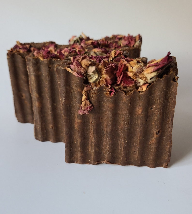 May include: Two brown bars of soap with dried rose petals embedded in the top. The soap has a ridged texture.