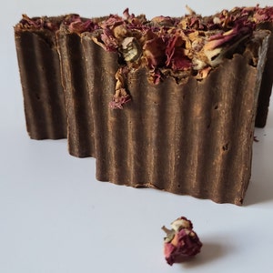 May include: Two brown bars of soap with dried rose petals on top. One bar is partially visible behind the other. A single dried rose petal is in the foreground.