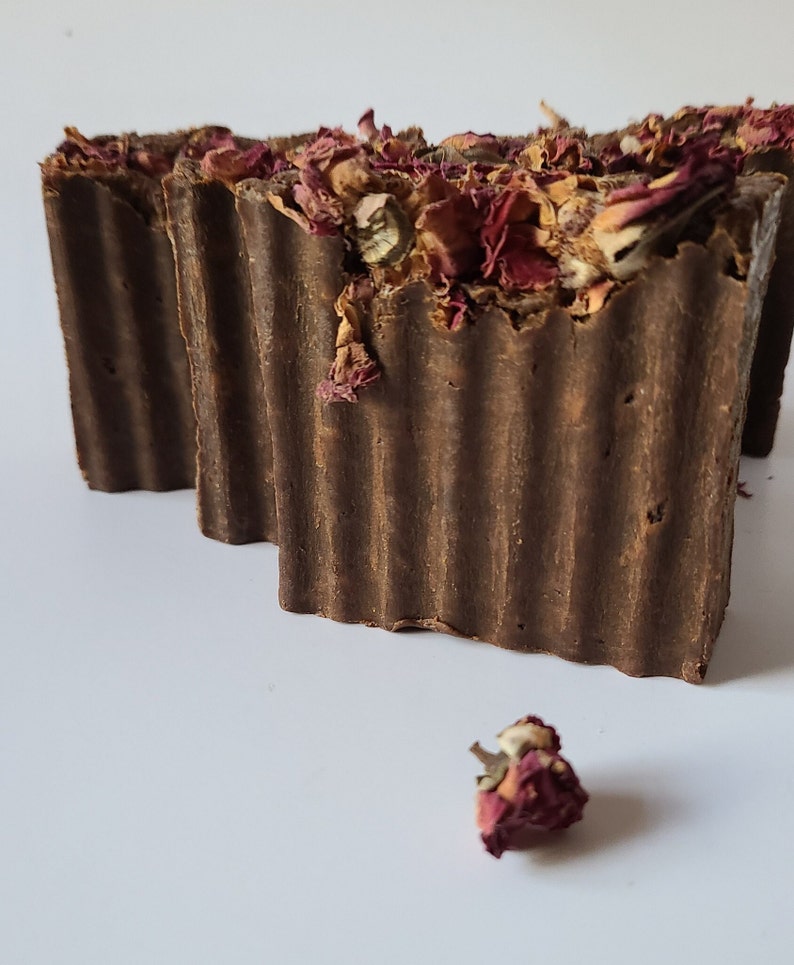 May include: Two brown bars of soap with dried rose petals on top. One bar is partially visible behind the other. A single dried rose petal is in the foreground.