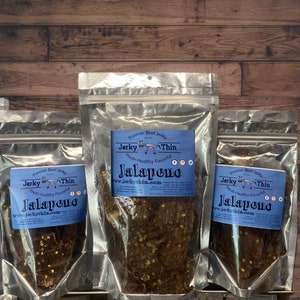 May include: Three clear plastic bags with resealable tops containing beef jerky. Each bag has a label with the text "Premier Beef Jerky" and "Jerky Thin" in a blue and white design. The text "Fresh-Healthy-Flavorful" is also on the label. The bags are labeled with the flavor "Jalapeno".
