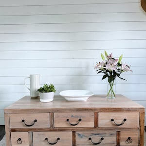 May include: A wooden coffee table with six drawers and black metal handles. The table is made of reclaimed wood and has a rustic finish. A white ceramic bowl, a white pitcher, and a potted succulent are on top of the table. A bouquet of pink lilies in a glass vase is on the table.