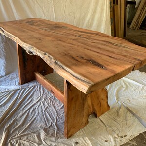 May include: A large, live edge wooden table with a natural wood finish. The table has a unique, irregular shape and is supported by a simple, modern base.