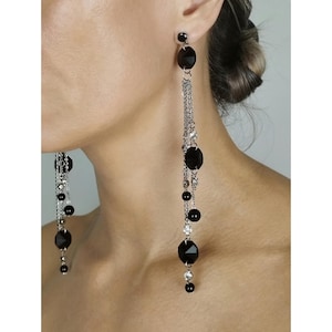 May include: Long, dangling earrings with black oval and round beads, and clear crystals. The earrings have a silver chain and a black stud at the top. The earrings are a statement piece.