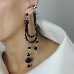May include: A black beaded earring with a silver ear cuff. The earring features multiple strands of black beads and dangling black crystal accents. The design includes a small stud and a chain that connects to the ear cuff.