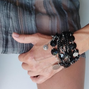 Könnte beinhalten: Ein schwarzes Armband mit mehreren Strängen und Anhängern. Das Armband hat facettierte Perlen, kleine runde Perlen und baumelnde silberfarbene Anhänger. Das Design umfasst eine Mischung aus Texturen und Formen, wodurch ein geschichteter Effekt entsteht.