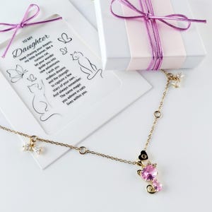 Puede incluir: Un collar de cadena dorada con un colgante en forma de gato rosa con cristales transparentes y pequeños dijes de lazo. El collar se presenta con una caja de regalo blanca y una tarjeta con el texto To My Daughter y un poema sobre gatos.