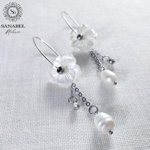Peut inclure: Boucles d'oreilles fleuries blanches avec des perles pendantes et des chaînes. Les boucles d'oreilles présentent un motif floral et des crochets argentés. Elles proviennent de la collection Sanabel Atelier.