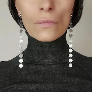 Può includere: Orecchini pendenti lunghi color argento con una serie di dischi circolari. Gli orecchini sono sospesi da un piccolo perno. La modella indossa un dolcevita nero.