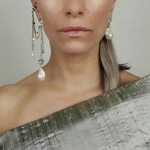 May include: Elegant silver earrings with dangling chains, crystals, and large, irregular white pearls. One earring is a stud with a single pearl, while the other features multiple strands. The earrings are a statement piece, adding a touch of luxury.