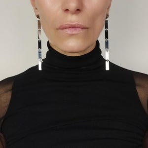 May include: Silver rectangular drop earrings. The earrings are composed of multiple rectangular pieces connected in a vertical line. The person in the image is wearing a black turtleneck.